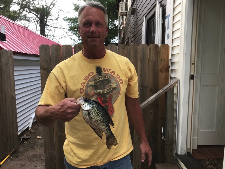 A man standing in front of his cabin, showing off a record sized bluegill fish he caught while fishing on the lake.